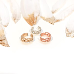 May include: Three adjustable toe rings in gold, silver, and rose gold. Each ring features a woven design. The rings are displayed on a white surface with blurred, feather-like accents in the background.