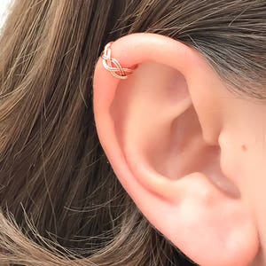 May include: A rose gold ear cuff featuring a delicate, interwoven wire design that wraps around the upper cartilage of the ear. The metal has a polished, metallic finish, creating a continuous, knotted pattern that sits securely on the ear edge.