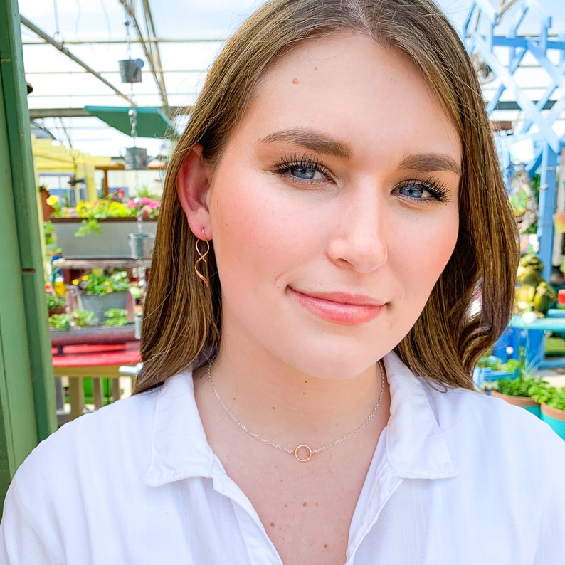 May include: A woman wearing a white shirt with a delicate gold necklace featuring a small circle pendant. She is also wearing a pair of gold earrings with a twisted design.