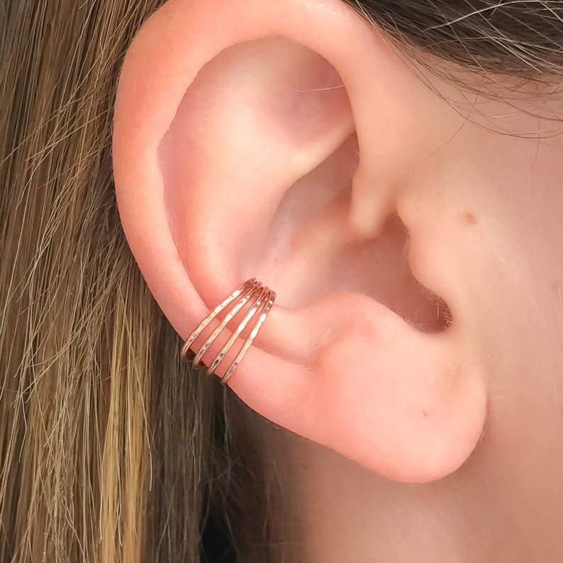 May include: A rose gold ear cuff with a hammered texture. The cuff features four bands that wrap around the ear. The jewelry is shown on a person's ear, with the ear in the background.