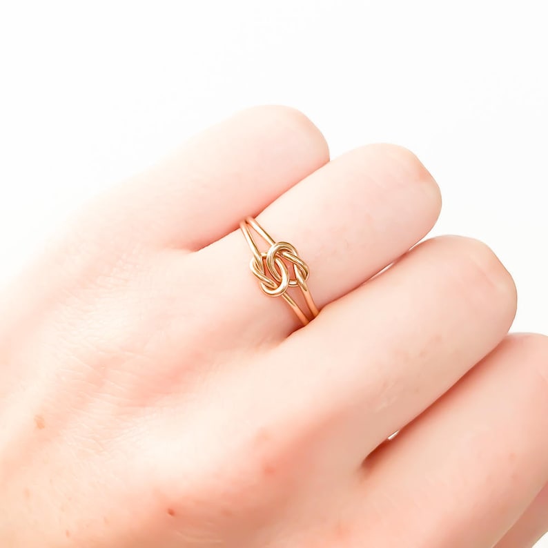 May include: A gold-toned ring featuring a central knot design. The band is constructed from thin, polished wire that loops into a symmetrical knot at the centre, creating a delicate and continuous circular shape that rests on the finger.