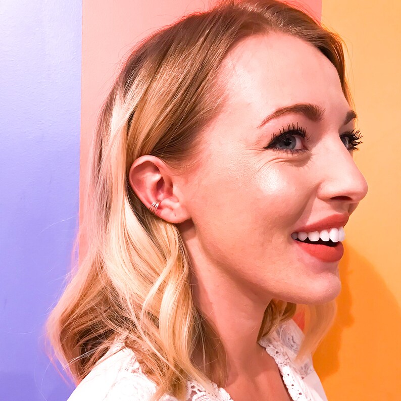 May include: A smiling person with blonde hair and a white lace top. The person is wearing a triple ear cuff on the left ear. The background is a gradient of pink, purple, and orange.