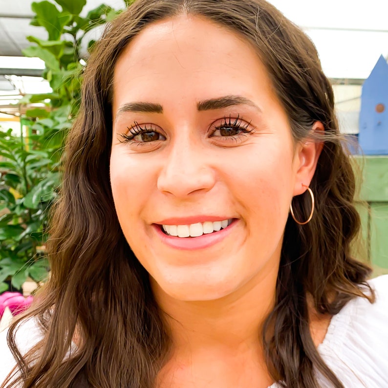 May include: A woman with long brown hair is smiling and wearing gold hoop earrings.