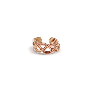 May include: A rose gold-colored toe ring with a braided design. The ring features an open design with a woven pattern across the top. The metal has a polished finish, reflecting light. The ring is set against a white background.