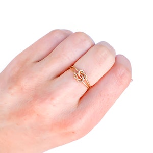 May include: A thin gold ring featuring a central knot design. The band is constructed from two delicate gold wires that cross and intertwine at the centre to form a small, symmetrical knot detail. The metal has a polished, reflective finish.