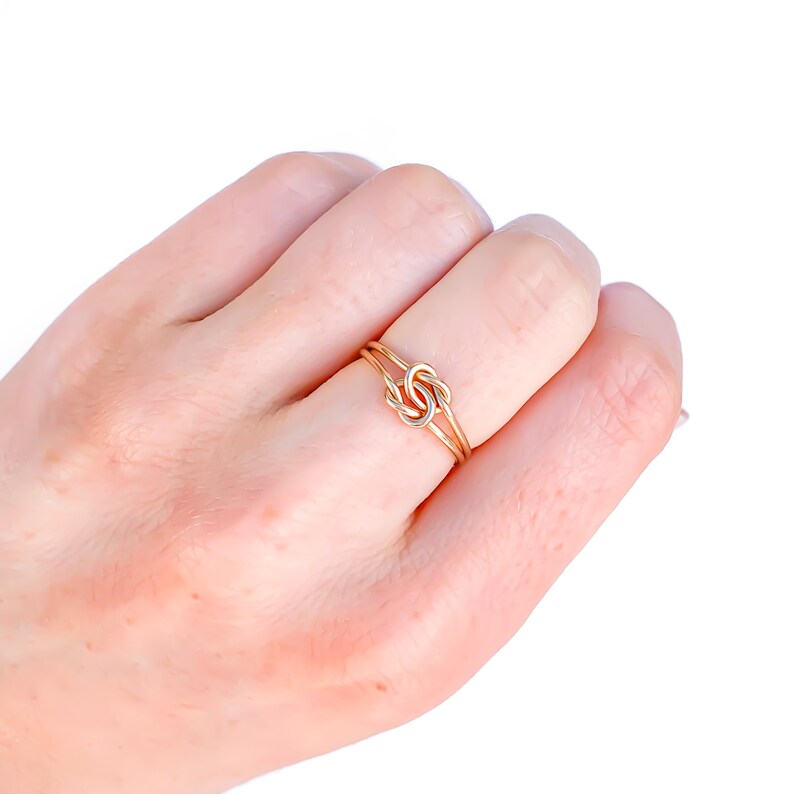 May include: A thin gold ring featuring a central knot design. The band is constructed from two delicate gold wires that cross and intertwine at the centre to form a small, symmetrical knot detail. The metal has a polished, reflective finish.
