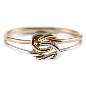 May include: A two-tone ring featuring a central knot design. The band is constructed from a thin, polished yellow gold wire and a thin, polished silver-colored wire that intertwine to form a singular, symmetrical knot at the front of the ring.