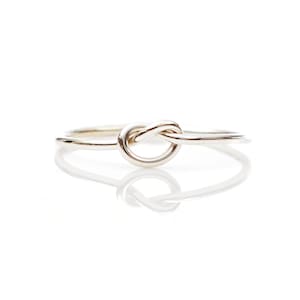 May include: A silver-colored ring with a knot design. The ring is simple and elegant, with a polished finish. The knot is the focal point, creating a unique and eye-catching piece of jewelry.