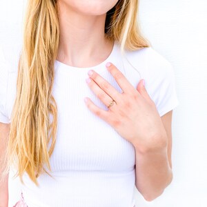 May include: A thin gold ring featuring a small, centred knot design. The band is smooth and circular, resting on a finger. The metal has a polished, reflective finish that catches the light, creating a simple and delicate appearance on the hand.