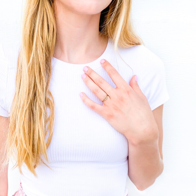 May include: A thin gold ring featuring a small, centred knot design. The band is smooth and circular, resting on a finger. The metal has a polished, reflective finish that catches the light, creating a simple and delicate appearance on the hand.
