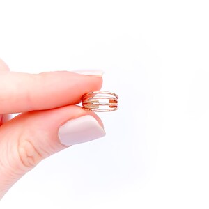 May include: A gold-colored ring with a cage-like design. The ring features multiple horizontal bars, creating an open, airy aesthetic. The ring is held between fingers against a white background.