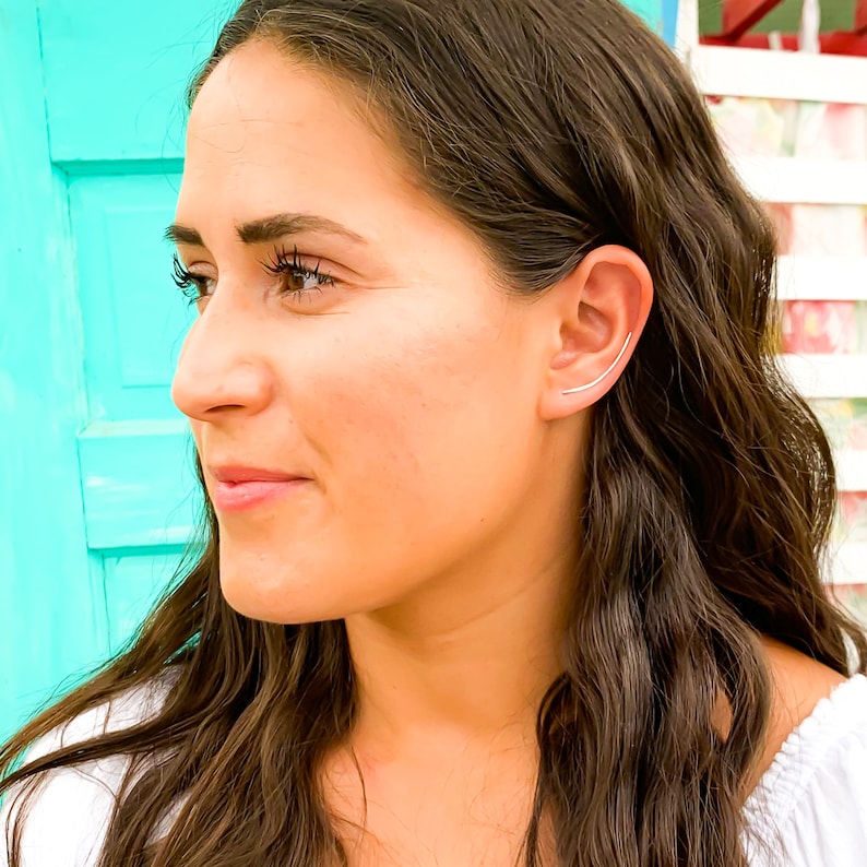 May include: A woman with long brown hair wearing a white shirt and a silver ear cuff. The ear cuff is a thin, curved bar that goes through the upper part of the ear.