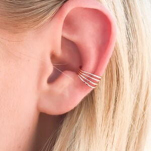 May include: A gold ear cuff with four stacked bands is worn on the ear. The jewelry piece is shown against the backdrop of a person's ear and blonde hair. The ear cuff is a simple, yet elegant accessory.