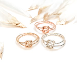 May include: Three rings made of thin wire, each featuring a central knot design. One ring is rose gold, one is yellow gold, and one is silver. The bands are constructed from double strands of wire that meet at the knot detail in the centre.