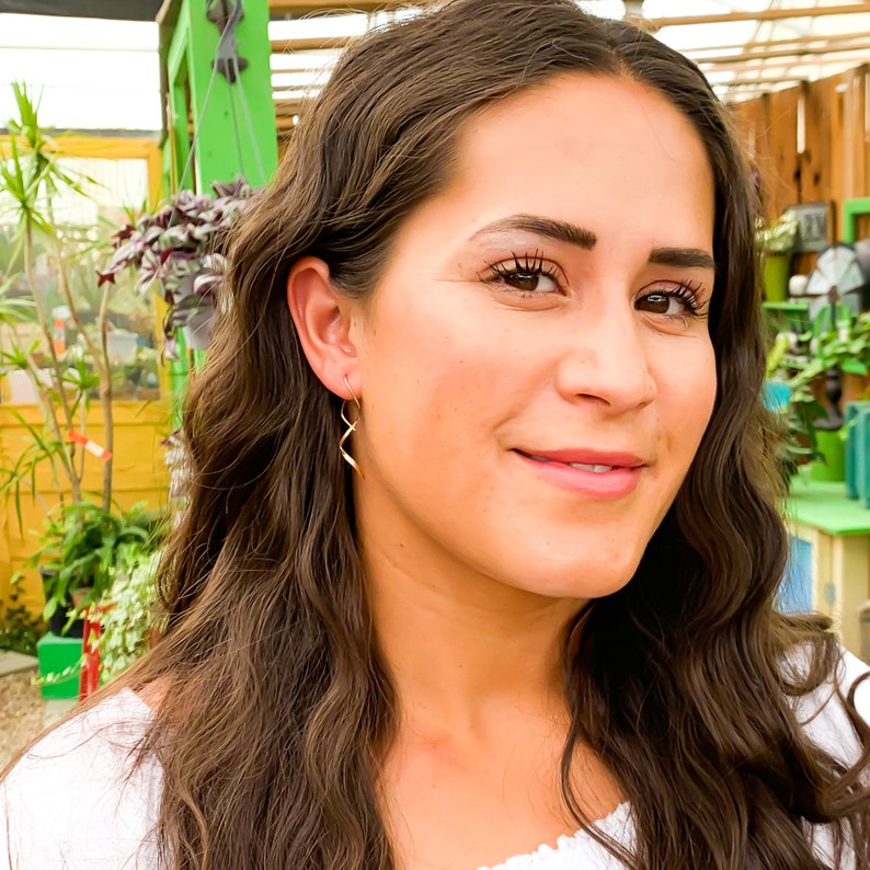 May include: A woman with long brown hair wearing a white shirt and gold earrings. The earrings are a twisted, thin, and long design.