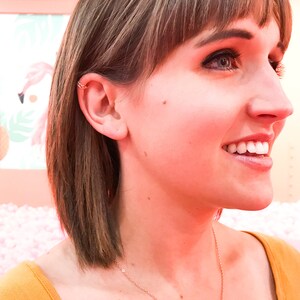May include: A gold-colored ear cuff is worn on the upper ear. The ear cuff has a woven design. The person in the image is smiling and wearing a gold necklace and a mustard-colored top. The background is a soft pink.