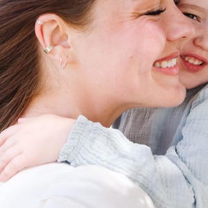 May include: A side profile of a smiling person with brown hair, wearing a gold ear cuff and small hoop earrings. Another person is embracing them, wearing a light blue shirt. The image is brightly lit.