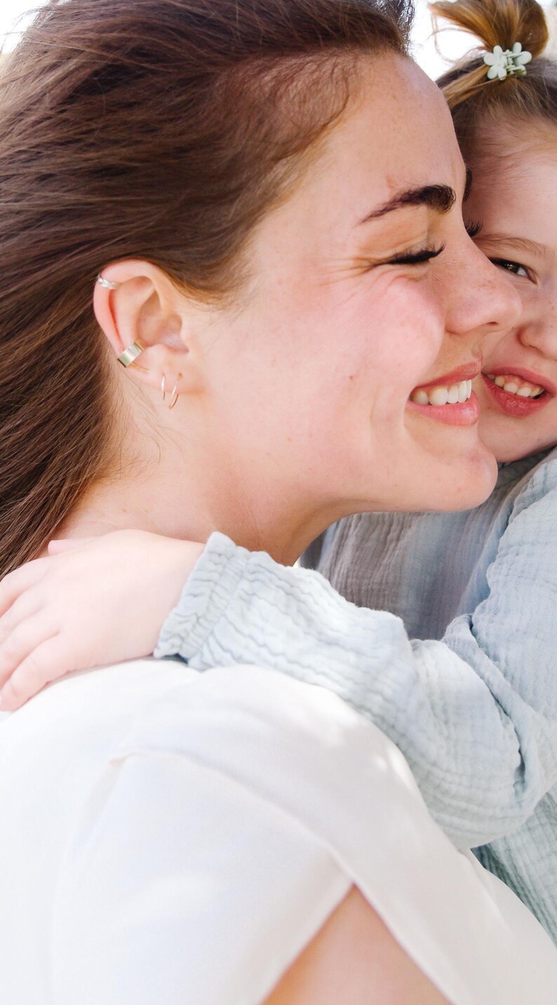 May include: A side profile of a smiling person with brown hair, wearing a gold ear cuff and small hoop earrings. Another person is embracing them, wearing a light blue shirt. The image is brightly lit.