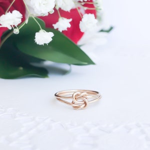 May include: A rose gold ring featuring a central knot design. The band is constructed from a single continuous wire that loops into a symmetrical knot at the front, creating a polished and smooth metallic finish with a warm pinkish hue.