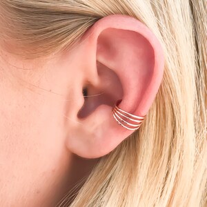 May include: Close-up of a gold ear cuff with four stacked bands. The cuff is worn on the upper ear cartilage. The background shows blonde hair and the ear's natural skin tone.