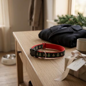 Puede incluir: Un collar para perros negro y rojo con un dise&ntilde;o de &aacute;rbol de Navidad. El collar est&aacute; sobre una mesa de madera con una chaqueta oscura, una taza y una caja de regalo. El collar tiene una anilla en D met&aacute;lica y una hebilla de pl&aacute;stico negra.