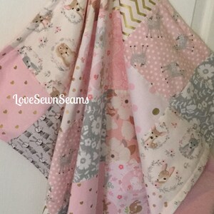 Puede incluir: Una manta de bebé de patchwork rosa y gris con un tema de animales del bosque. La manta presenta ciervos, conejos y patrones florales. El texto "Love Sewn Seams" está impreso en la manta.