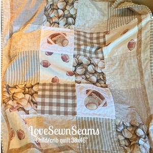 May include: A patchwork quilt featuring a sports theme with baseball and football patterns in shades of tan, brown, and white. The quilt includes checkered and striped fabric squares, with text reading LoveSewnSeams Child/crib quilt 38x46 inches.