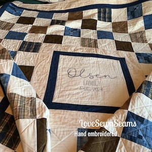 May include: Hand-embroidered quilt with a patchwork design in shades of blue, brown, and cream. The center features the name "Olsen Family" and "Est. 2024". The quilt is made by LoveSewnSeams.