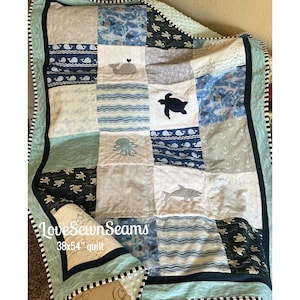 May include: A patchwork quilt featuring various sea life patterns and appliqués including a whale, turtle, octopus, and shark. The quilt displays blue, white, and grey tones with a black and white striped border and text reading LoveSewnSeams 38x54 quilt.
