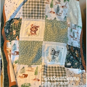 Könnte beinhalten: Ein Patchwork-Kuscheldecke mit Waldthema. Die Decke zeigt Quadrate mit Illustrationen von Bären, Füchsen und Hirschen sowie grüne Vichy- und gepunktete Muster. Die Decke misst 96.5x96.5 cm.