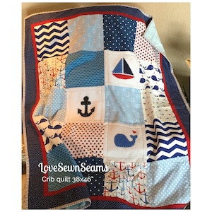 May include: A blue and red crib quilt with a nautical theme. The quilt features anchors, whales, sailboats, and zig-zag patterns. The quilt is 38 inches by 46 inches. "Love Sewn Seams" is written on the quilt.