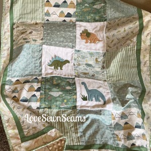 Może przedstawiać: Pikowana narzuta typu patchwork wykonana z różnych tkanin w kolorze szałwiowej zieleni, bieli i błękitu, ozdobiona wzorami dinozaurów i gór. Trzy białe kwadratowe panele przedstawiają wyhaftowane dinozaury: triceratopsa, stegozaura i zauropoda.