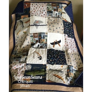 Op de afbeelding: Een patchwork wiegdeken met stofvierkanten met een visthema. De quilt bevat afbeeldingen van vissen, hengels, kano's en de tekst "Catch of the Day". De quilt meet 96,5x117 cm.