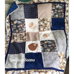 Op de afbeelding: Een patchworkdeken met een baseballthema. De deken is gemaakt van verschillende stoffen, waaronder blauw ruit, bruine strepen en wit met bedrukte baseballs. De deken heeft ook aangebrachte baseballs, een honkbalhandschoen en de tekst "LoveSewnSeams".