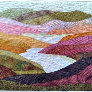 Puede incluir: Un paisaje acolchado con colinas onduladas en tonos rosa, naranja, verde y marr&oacute;n. El edred&oacute;n tiene un borde blanco y representa un r&iacute;o o valle. Las costuras a&ntilde;aden textura y detalle a la obra de arte.