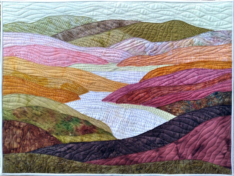 Puede incluir: Un paisaje acolchado con colinas onduladas en tonos rosa, naranja, verde y marr&oacute;n. El edred&oacute;n tiene un borde blanco y representa un r&iacute;o o valle. Las costuras a&ntilde;aden textura y detalle a la obra de arte.