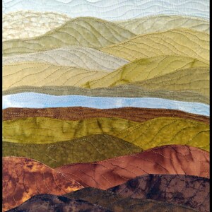 May include: A quilted wall art piece depicting a landscape with rolling hills in shades of brown, green, and blue. The hills are stitched together with a wavy pattern, creating a textured effect.