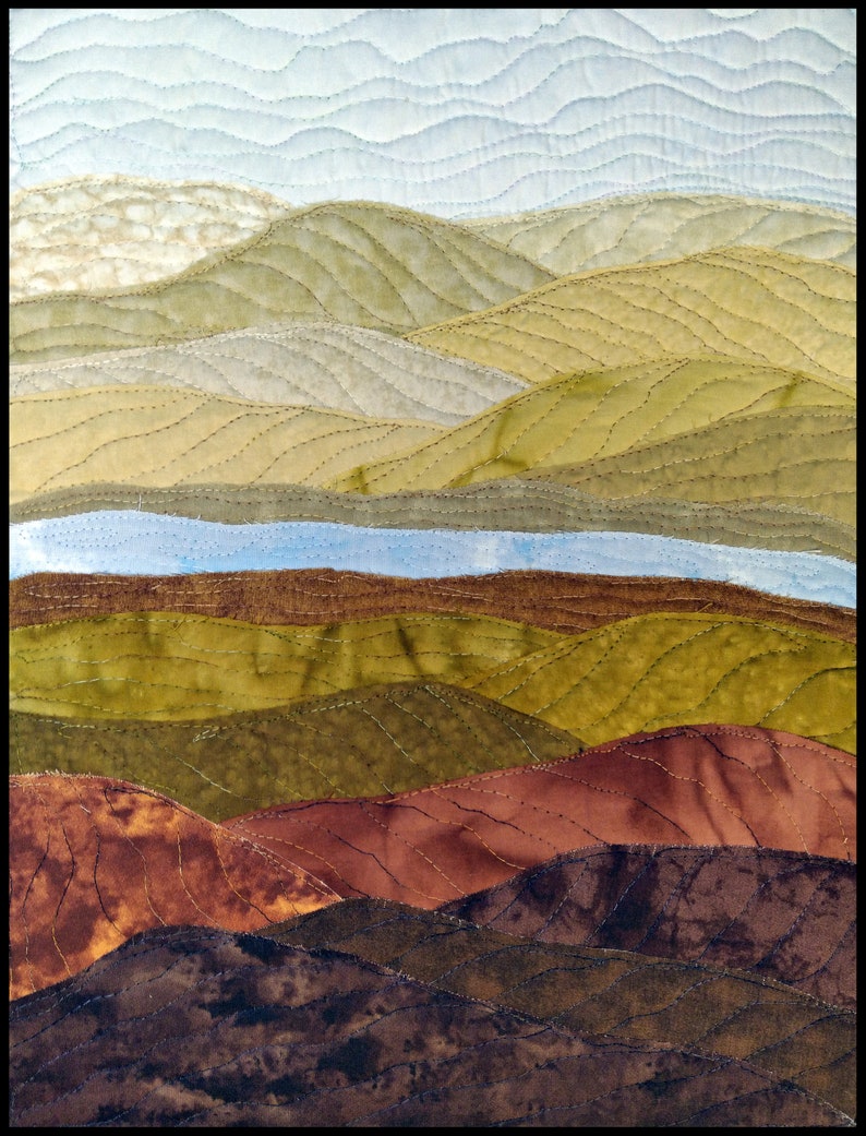 May include: A quilted wall art piece depicting a landscape with rolling hills in shades of brown, green, and blue. The hills are stitched together with a wavy pattern, creating a textured effect.
