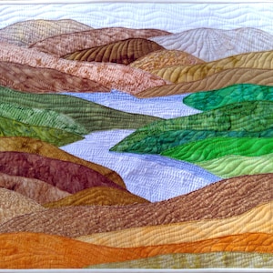 Puede incluir: Un paisaje acolchado que representa colinas y un r&iacute;o. La obra de arte presenta una variedad de tonos tierra, incluyendo tonos de marr&oacute;n, verde y naranja. El r&iacute;o es de color azul claro y el cielo es blanco p&aacute;lido. El edred&oacute;n est&aacute; enmarcado con un borde blanco.