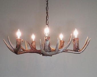 Può includere: Un lampadario rustico a corna con cinque lampadine. Il lampadario è fatto di corna dall'aspetto naturale e ha una catena per appenderlo. Le lampadine sono accese, proiettando un bagliore caldo.