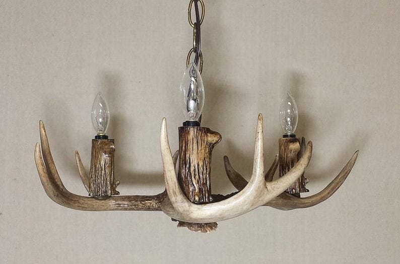 May include: A rustic chandelier made from antlers with three light bulbs. The antlers are a natural light brown colour and the chandelier is hanging from a chain.
