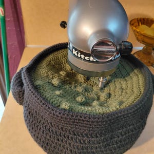 May include: A silver KitchenAid stand mixer head positioned above a dark gray crocheted bowl cover with a textured olive green center. The mixer features a metallic finish and black control knobs, while the cover has a dense, yarn-based construction.