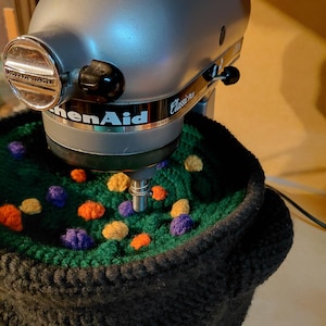 May include: A silver KitchenAid Classic Plus stand mixer positioned above a black crocheted cauldron cover. The cover features a dark green textured interior with small, scattered orange, yellow, and purple crocheted spheres resembling bubbles.