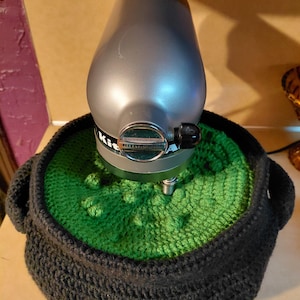 May include: A silver KitchenAid stand mixer base is partially covered by a dark charcoal gray crocheted cover with a bright green textured top featuring raised bobble stitches, designed to fit around the mixer's central housing.