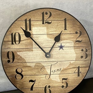 May include: A round wooden clock with a brown and white woodgrain design. The clock face features a silhouette of the state of Texas with a blue star in the center. The clock hands are black and the numbers are black on a white background. The clock is on a black stand. The text "Texas" is written in cursive on the clock face.