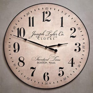 May include: A round wall clock with a beige, textured face and a thin black frame. It features large black Arabic numerals, black hands, and the text Joseph Tyler Co. CLOCKS, Standard Time, and BOSTON, MASS printed in black on the clock face.