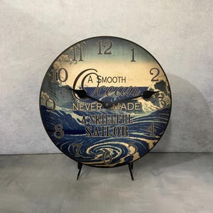 May include: A round wooden clock with a blue and white ocean wave design. The clock face has black Roman numeral hour markers and black hands. The clock has a black stand and the text "A smooth ocean never made a skillful sailor".