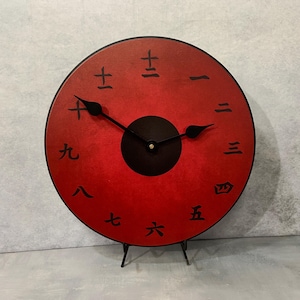 May include: A red clock with black hands and black numbers in Japanese characters. The clock is on a black stand.