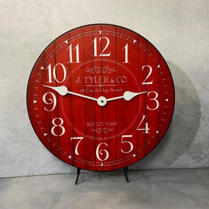 May include: Red round clock with white Roman numeral markings and black hands. The clock face has a distressed woodgrain design and features the text "J. TYLER & CO. 82 Cambridge Street BOSTON MASS".
