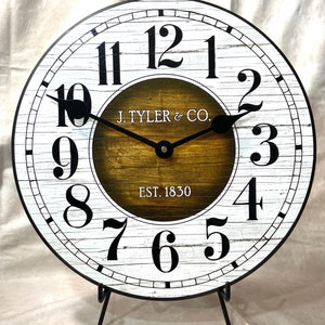 May include: A white and black farmhouse style clock with black hands and a brown woodgrain center. The clock face reads "J. TYLER & CO. EST. 1830".